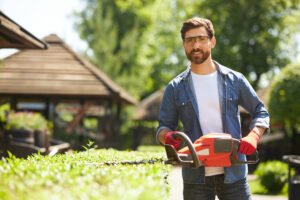 Happy, attractive male gardener in glasses posing, while trimming bush with electric hedge clippers. Portrait of smiling handyman with beard shaping boxwood bush at backyard. Concept of landscaping.